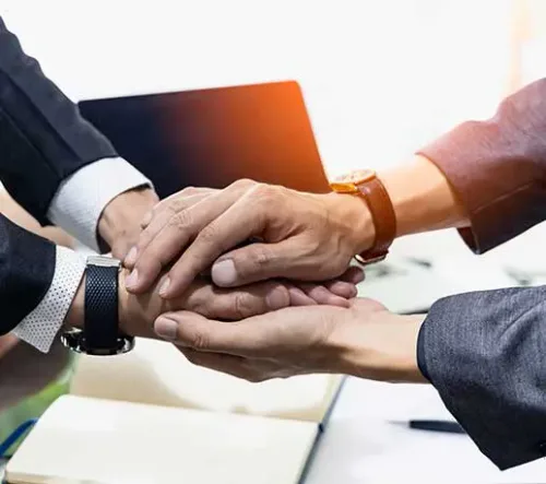 Three people in business suits place their hands together in a gesture of teamwork or agreement, with a laptop and notebook visible in the background.