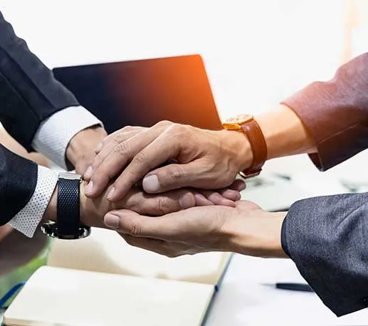 Three people in business suits place their hands together in a gesture of teamwork or agreement, with a laptop and notebook visible in the background.